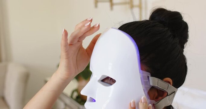 Woman is lifting LED mask onto face on bed, aligning cutouts and securing strap for therapy