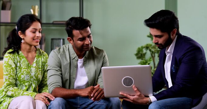 Indian couple meeting agent at home with laptop during investment discussion with bank representative reviewing financial plans while sitting on sofa in cozy modern living room, home consultation