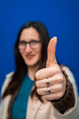 Smiling woman with glasses giving a thumbs up gesture, indicating agreement, success, and positivity on a blue background