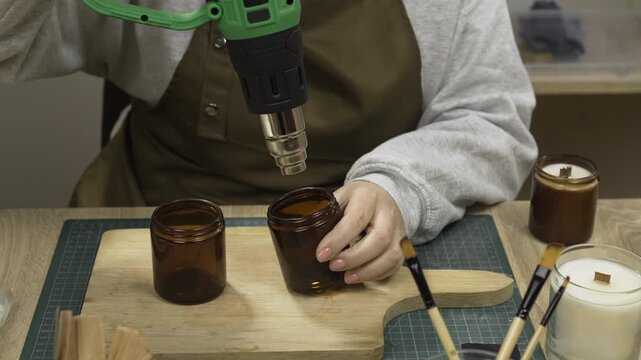 Close-up of female hands heating glass jars while making homemade wax candles in a workshop using a heat gun.