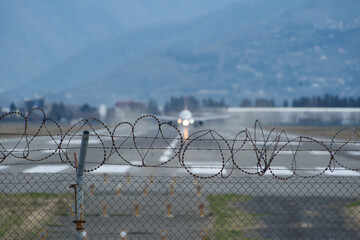 Barbed wire fence by the airport, blurry plane taking off and mountains beyond