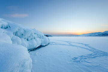 Frozen ice formations at Winter sunrise with footprints zigzagging around the cliff. Smooth snow surface create a minimalistic arctic scenery with soft pastel sky.