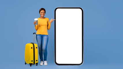 A person stands with a yellow suitcase while holding a cup of coffee. They point towards a large empty phone screen. The background is a solid light blue color. © Prostock-studio
