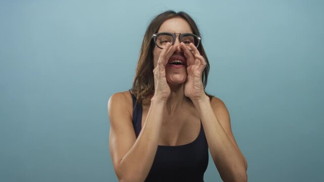 Woman wearing glasses calls out with hands cupped around her mouth in a blue studio; excitement.
