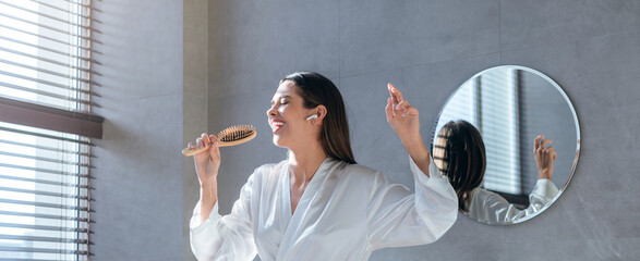 Woman stands in a bright bathroom, singing while brushing her hair with a wooden brush. Sunlight shines through blinds as she moves with joy. The mirror reflects her energy. © Prostock-studio