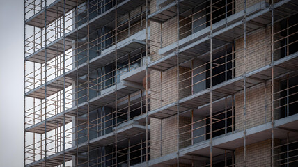 Fototapeta premium Scaffolding surrounds a partially constructed high rise building facade featuring unfinished windows and light colored brickwork against the sky.