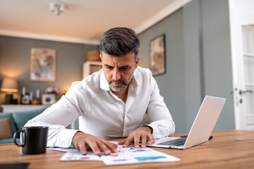 Fototapeta premium Serious businessman reviewing data on paper charts at office desk with laptop. Business and finance concept. Focused man checking financial reports and statistics while working in home office.