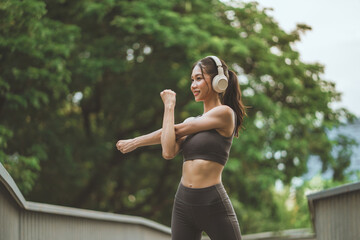 Smiling Asian woman athlete wearing headphones in sports bra stretching her arms before a workout in a green park. Healthy lifestyle fitness and outdoor exercise concept.