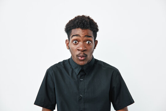 Young African American man with dark skin and curly hair wearing black shirt looking surprised with wide eyes and open mouth on white background in studio portrait.
