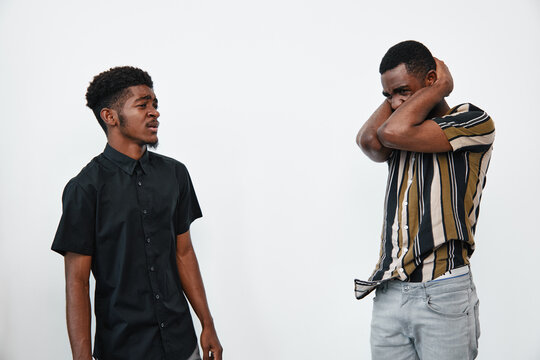 Two young black men with dark skin in casual clothes having a tense conversation in studio. One man in black shirt looks serious, other in striped shirt covers face with arms.