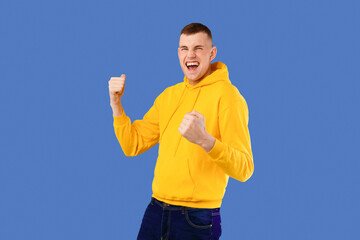 Naklejka premium Overjoyed caucasian man shouting and shaking fists, celebrating big success over blue studio background, free space. Celebration of victory, joy emotion concept