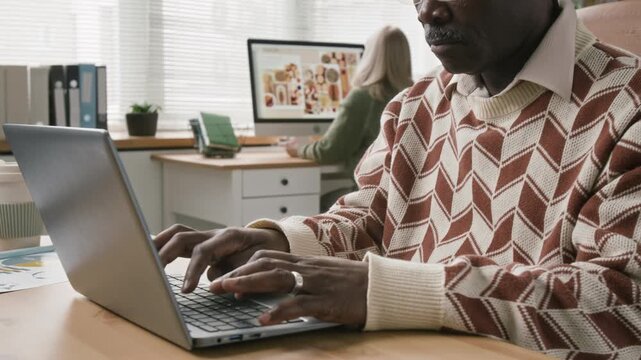 Tilt down shot of senior Black male office manager fitting eyeglasses then typing email on laptop while working at desk in modern open space with colleagues