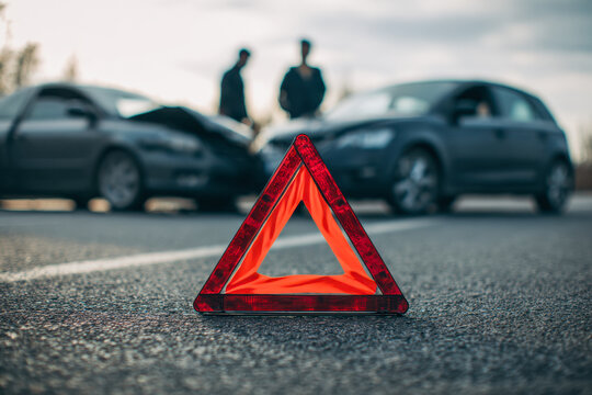 Reflective emergency warning triangle placed on road in front of two damaged vehicles with two people discussing car accident aftermath on cloudy day