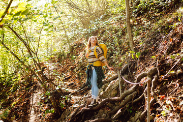 Fototapeta premium A female hiker with a backpack walks along a path in a rocky forest. A young woman on a hike, enjoying the sunset and the adventure in the mountains. Active lifestyle. Concept: recreation, hiking.
