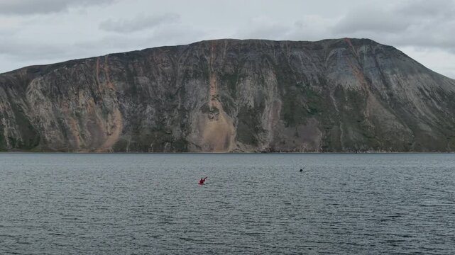 Drone footage shows a rugged sea cliff rising above dark water near Penkigney Bay. Slow pan reveals the remote Chukotka coast of under heavy clouds. Tiny boats dot the Bering Strait