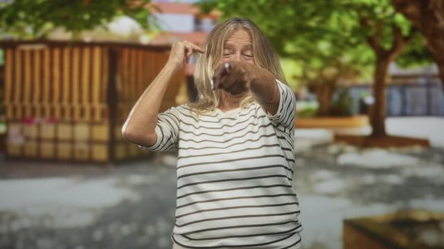 Senior woman smiling and pointing finger with right hand at camera on street near wooden kiosk wearing striped shirt; playful challenge.