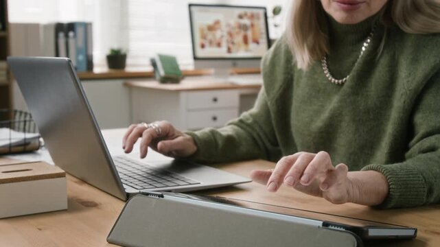 Cropped shot of senior female office manager writing emails on laptop then checking information on digital tablet while multitasking between devices at working desk in modern open space