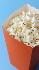 Freshly popped white popcorn in a vibrant orange box on a bright blue background. Movie night snack for entertainment and relaxation.