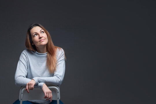 Woman sits on chair and looks up with calm expression in studio setting during indoor photo session in soft lighting conditions