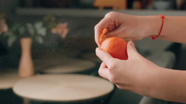 Woman peeling fresh ripe tangerine in slow motion with zest spraying, creating citrus mist. Close-up of female hands holding juicy organic fruit for healthy snack