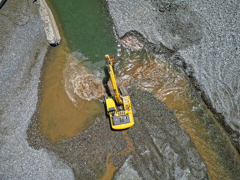 A yellow excavator is working on a gravel bank at the edge of a river delta, reshaping the shoreline. Brown sediment clouds the water, showing active digging in a coastal or riverbed environment.

