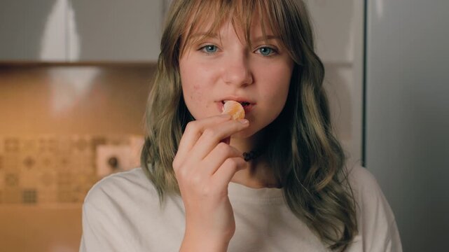 Close-up portrait of teenager girl with blue eyes and bangs eating juicy mandarin orange slice in modern kitchen while look directly at camera.