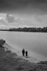 Caucasian man and young girl in dark coats holding hands on sandy river shore, looking away. Wide river, trees, cloudy sky. Black and white image with copy space. Concepts: family, childhood.