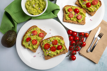 Overhead view of avocado toast topped with fresh cherry tomatoes and red onion on whole grain bread, served on white and red plates with fresh ingredients. This clean and colorful composition represen