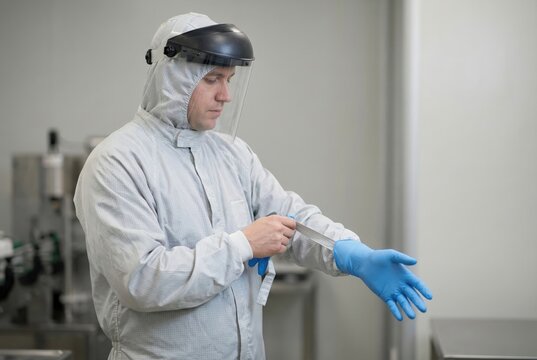 A male worker in a white protective suit and face shield is putting on blue disposable gloves in a sterile laboratory or factory environment.