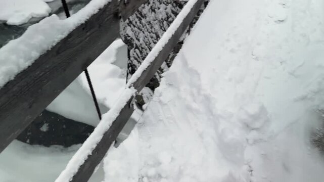 Man clearing deep snow with a shovel on a narrow suspension footbridge in winter Carpathians, Ukraine. Difficult winter maintenance, rural life and dangerous work in mountain forest landscape.