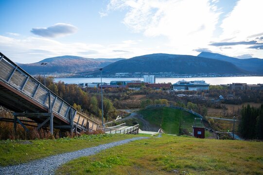 Modern ski jump facility with scenic walkway overlooking Tromso city and fjord in northern Norway. Dramatic mountain backdrop with autumn colors creates stunning Nordic landscape vista.