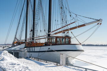 Fototapeta premium Large historic schooner docked at a snowy pier, its rigging and masts prominent against a clear blue sky, with a wide expanse of ice covering the surrounding water