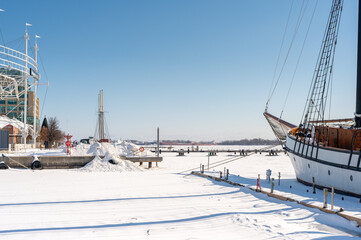 Fototapeta premium Sailboat docked on a frozen lake with snow covered piers and distant buildings under a clear blue sky, depicting the cold winter season at the city waterfront