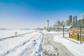 Fototapeta premium Snow covering a pedestrian path and the frozen surface of lake ontario, with the harbourfront centre building and the toronto city skyline under a clear blue winter sky
