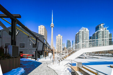 Fototapeta premium Toronto cityscape showcasing a prominent landmark tower rising above urban buildings and a waterfront area covered in snow on a bright, clear blue sky day