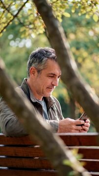 Smiling mature man using smartphone on park bench framed by tree branches for authentic digital connection and mobile app marketing in natural outdoor setting