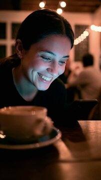 Smiling young woman using smartphone in cozy night cafe with warm string lights and coffee cup for authentic digital connection and mobile social media engagement concept with cool screen glow
