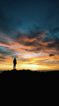 Lone silhouette standing on rocky mountain peak at sunset with dramatic orange and blue sky for adventure travel, mystic outlands, and poetcore mindfulness concept