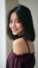 Young woman with dark hair and purple top smiling and looking over shoulder, indoor portrait