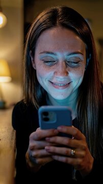 Smiling young woman with freckles using smartphone in cozy night cafe with warm string lights and coffee cup for authentic connectioneering and urban social media concept