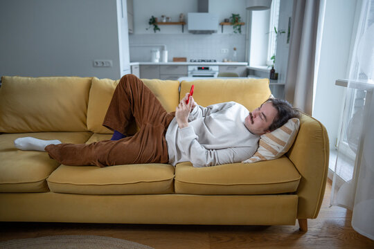 Lazy bored man lying on sofa scrolling phone with social media, procrastinating, lack of motivation. Digital addiction of male rest on sofa using smartphone, avoiding responsibilities, delaying tasks.