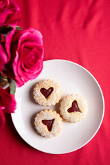 Over head view and Close-up of a heart-shaped jam-filled cookie dusted with powdered sugar and served on a white plate. The soft focus background and warm red tones create a romantic, cozy mood, makin