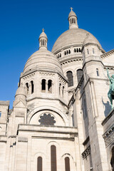 Obraz premium Low angle view of the white domes and intricate stone architecture of the Sacre Coeur Basilica in Montmartre, Paris, under a clear blue sky, famous French religious landmark and tourist attraction