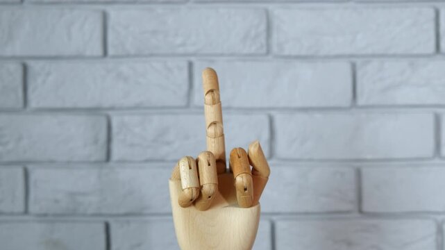 Wooden mannequin hand making obscene gesture. Articulated wooden hand model rising from the bottom of the frame to show an offensive middle finger gesture against a gray brick wall background