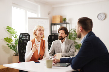 Fototapeta premium A group of three professionals discuss ideas in a meeting room. They share thoughts while seated at a table with plants in the background. Each person has a cup and materials.