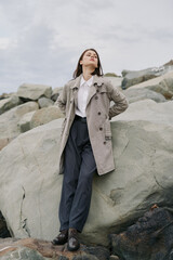 Young woman in long grey coat stands confidently on large rocks under cloudy sky. Fashion portrait of stylish model in casual outfit outdoors with natural background and moody atmosphere. © SHOTPRIME STUDIO