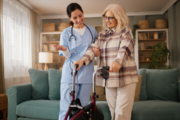A home health nurse helps an elderly woman with a walker in a living room. The two are talking as the nurse offers support during an afternoon home care visit.
