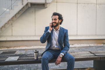Obraz premium Businessman talking on phone while sitting on bench outside office