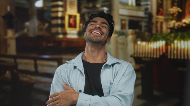 Man in denim shirt smiling with hand to cheek beside a worn pew and row of candles in building; serenity.
