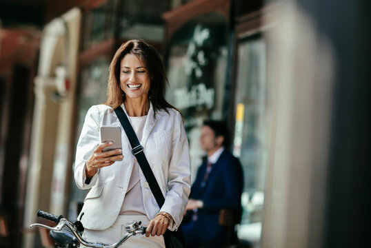Smiling businesswoman using smartphone with bicycle on city street